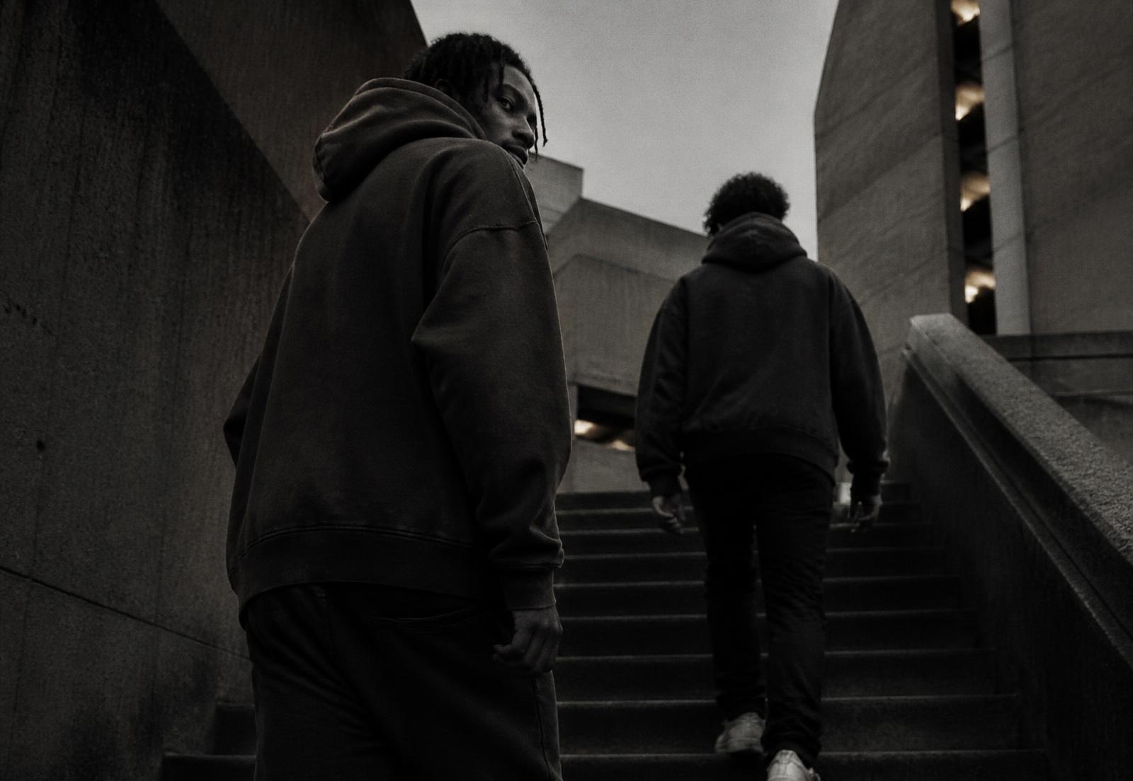 Two friends in oversized hoodies climbing a concrete stairway between brutalist buildings at dusk